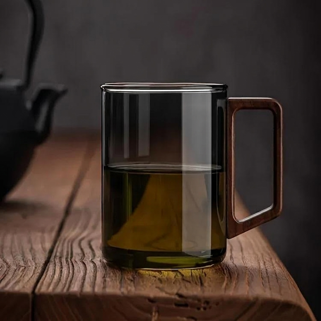 Caneca de vidro borossilicato fumado com pega retangular de madeira maciça de nogueira, preenchida com chá de tom âmbar, pousada sobre uma mesa de madeira rústica texturada em ambiente de luz suave e fundo escuro minimalista.