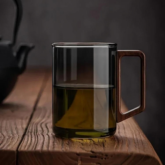 Caneca de vidro borossilicato fumado com pega retangular de madeira maciça de nogueira, preenchida com chá de tom âmbar, pousada sobre uma mesa de madeira rústica texturada em ambiente de luz suave e fundo escuro minimalista.