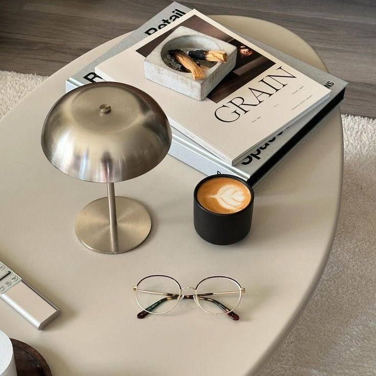 Um candeeiro de mesa prateado, moderno e com acabamento reflexivo em forma de cogumelo, posicionado sobre uma mesa de centro estilizada ao lado de um livro de café intitulado 'GRAIN', uma chávena de café escura e um par de óculos de leitura finos.