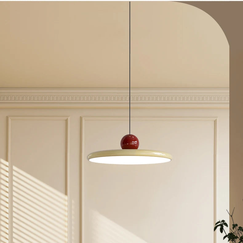 Candeeiro de suspensão minimalista em tom creme com uma esfera bordeaux no topo, suspenso sobre uma mesa redonda branca. Ao fundo, destaca-se um arco arquitetónico elegante e uma janela com persianas por onde entra uma luz solar suave, iluminando cadeiras de design em madeira e palhinha num ambiente de estética nórdica e sofisticada.