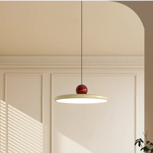 Candeeiro de suspensão minimalista em tom creme com uma esfera bordeaux no topo, suspenso sobre uma mesa redonda branca. Ao fundo, destaca-se um arco arquitetónico elegante e uma janela com persianas por onde entra uma luz solar suave, iluminando cadeiras de design em madeira e palhinha num ambiente de estética nórdica e sofisticada.