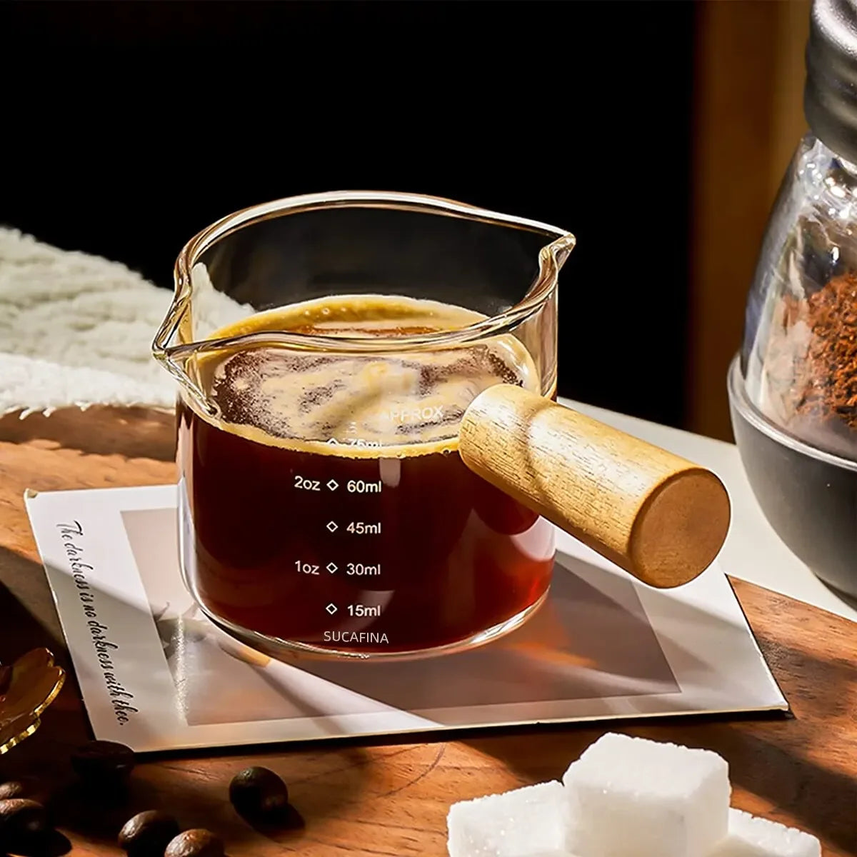 Jarra doseadora em vidro borossilicato com escala de medição e pega de madeira vertendo café num copo com leite, ambiente KURA Vibe