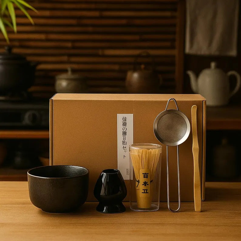 Conjunto de cerimónia tradicional de matcha com seis peças dispostas em linha, incluindo uma taça chawan de cerâmica escura texturada, um suporte cerâmico azul-claro, um batedor chasen de bambu, um coador metálico e uma colher chashaku de bambu, com a caixa de cartão de envio ao fundo.
